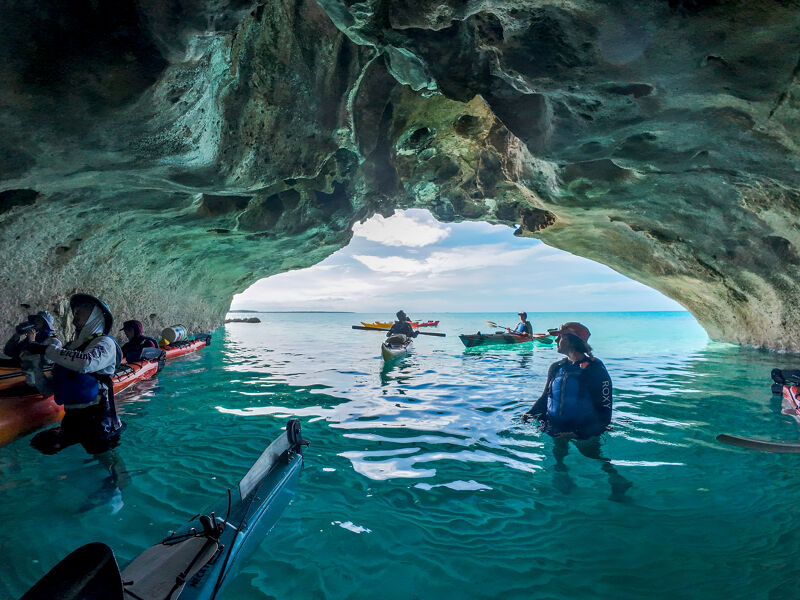 The image shows a group of people kayaking in a clear, turquoise sea, as seen from inside a cave. The cave's opening frames the kayakers and the horizon, creating a picturesque view. The water is calm, and the sky is visible in the distance. One person is standing in the water near the cave entrance, holding a paddle.